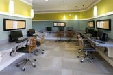 This single-floor business center is a thoughtfully designed shared workspace that balances functionality with a lively aesthetic. The room's most striking visual feature is its bold two-tone wall treatment, pairing chartreuse yellow on the upper walls with soft seafoam blue on the lower portion, separated by a crisp white chair rail — creating an energetic yet professional atmosphere. Crown molding along the ceiling adds a refined architectural detail.

Workstations line the walls in a U-shaped configuration, each equipped with desktop computers, keyboards, and monitors, accommodating multiple users simultaneously. Framed cork bulletin boards are mounted above the workstations for informational displays. Stylish orange lattice-back chairs on casters complement the colorful décor throughout the space.

A dedicated alcove houses an additional workstation alongside a printer, offering a quieter area for focused tasks. Pendant lighting with cylindrical glass shades hangs from the ceiling, supplemented by recessed lighting, ensuring well-distributed illumination throughout.

A seating nook with patterned accent chairs and a floor lamp is positioned beside a large multi-pane window, which floods the space with natural light and offers views of the surrounding streetscape and neighborhood. Patterned carpet tile flooring completes the contemporary, welcoming environment.