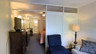 Located at 1865 Lakeland Dr in Jackson, Mississippi, this single-floor space offers a compact and functional layout designed to maximize utility. The interior features a semi-open floor plan where the living, sleeping, and kitchen areas flow into one another, separated by a partial partition wall with transom-style upper panels that allow light to pass between spaces, maintaining a sense of openness while providing visual division.

The living area includes a navy blue upholstered lounge chair, a floor lamp, and a side table, creating a modest seating nook. Adjacent to this is a sleeping area with a bed dressed in warm tones and a dark wood multi-drawer dresser, providing ample storage. A mirror is positioned nearby, adding depth to the space.

Toward the rear, the kitchen is visible and includes white cabinetry, a refrigerator, and overhead lighting. The kitchen area benefits from a window that brings in natural light. Louvered closet doors line one wall, offering additional storage. Carpeted flooring runs throughout, and ceiling-mounted light fixtures illuminate the space. The overall ambiance is functional and straightforward.
