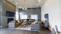 Located at 900 Reisling Lane in West Chester, PA, this well-appointed single-floor space offers a refined and welcoming environment designed with contemporary sensibilities. The centerpiece of the space is a generously proportioned lounge area that immediately impresses with its soaring coffered ceilings, which lend a sense of grandeur and architectural elegance. Recessed lighting and a modern chandelier work together to cast a warm, even glow throughout the room, enhancing the sophisticated atmosphere. A dramatic slate-blue accent wall serves as a bold focal point, complemented by framed abstract artwork and decorative wall accents that add personality and depth. Large, plush sectional sofas in warm neutral tones are arranged to encourage both conversation and relaxation, anchored by a layered area rug over rich, dark hardwood-style flooring. A flat-screen television and a media console on the opposite wall provide additional functionality. Natural light filters in through windows along the perimeter, brightening the space and connecting the interior to the outdoors. The overall ambiance is polished yet comfortable, striking a balance between upscale design and everyday livability, making this a standout destination in the West Chester area.