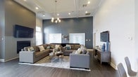 Located at 900 Reisling Lane in West Chester, PA, this well-appointed single-floor space offers a refined and welcoming environment designed with contemporary sensibilities. The centerpiece of the space is a generously proportioned lounge area that immediately impresses with its soaring coffered ceilings, which lend a sense of grandeur and architectural elegance. Recessed lighting and a modern chandelier work together to cast a warm, even glow throughout the room, enhancing the sophisticated atmosphere. A dramatic slate-blue accent wall serves as a bold focal point, complemented by framed abstract artwork and decorative wall accents that add personality and depth. Large, plush sectional sofas in warm neutral tones are arranged to encourage both conversation and relaxation, anchored by a layered area rug over rich, dark hardwood-style flooring. A flat-screen television and a media console on the opposite wall provide additional functionality. Natural light filters in through windows along the perimeter, brightening the space and connecting the interior to the outdoors. The overall ambiance is polished yet comfortable, striking a balance between upscale design and everyday livability, making this a standout destination in the West Chester area.