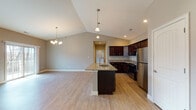 Located at 48 Bavarian Wy in Altamont, NY, this single-floor residence combines modern finishes with a thoughtfully designed open layout. The heart of the home is the open-concept kitchen and living area, where soaring vaulted ceilings create an impressive sense of volume and airiness. The kitchen is outfitted with rich dark cabinetry, granite countertops, a center island with a built-in sink, and a full suite of stainless steel appliances including a range, microwave, and refrigerator. Light wood-tone flooring runs throughout, complementing the neutral gray walls and white trim. A chandelier and pendant lighting add warmth to the dining and kitchen areas. The living space flows seamlessly toward a dedicated laundry closet with a stacked washer and dryer. Bedrooms are generously proportioned, with large windows that invite natural light and views of the surrounding landscape. The primary bathroom features a dual-sink vanity with granite counters, dark wood cabinetry, oval mirrors, and a walk-in shower. A secondary bathroom mirrors the same refined aesthetic with a single vanity, granite top, and a tub-shower combination. A walk-in closet off the primary bedroom adds practical storage. Recessed lighting and ceiling fans throughout complete this well-appointed home.