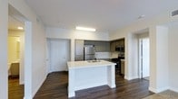 This single-floor space showcases a thoughtfully designed open-concept layout, with the kitchen serving as the focal point of the living area. A prominent white kitchen island anchors the space, offering a built-in sink and generous counter space, making it ideal for meal preparation and casual dining. The island's clean white finish contrasts attractively with the dark hardwood-style flooring that extends throughout the unit.

The kitchen is outfitted with stainless steel appliances, including a full-size refrigerator, a gas range, and an over-the-range microwave. Gray cabinetry lines the perimeter walls, providing ample storage, while light-toned countertops complement the overall neutral color palette. A flush-mounted ceiling light fixture provides direct illumination over the kitchen workspace.

The open floor plan allows the kitchen to flow seamlessly into the adjacent living area, creating a connected and versatile space. White walls and white trim throughout contribute to a bright, clean aesthetic. A white paneled door, visible from the kitchen, leads to an additional room or entryway, adding to the unit's functional layout. The overall ambiance is modern, minimal, and move-in ready.