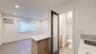 Located at 1225 N El Centro Ave in the heart of Hollywood, Los Angeles, this thoughtfully designed single-floor unit offers a clean, modern aesthetic with efficient use of space. The open-concept layout centers around a stylish kitchen island with wood-grain cabinetry and a crisp white countertop, providing both storage and a functional surface. The kitchen is well-appointed with upper wood-toned cabinets, a stainless steel microwave and dishwasher, a subway tile backsplash, and a sink — all framed by recessed lighting that keeps the space bright and welcoming. Large-format neutral floor tiles run throughout the main area, lending a cohesive and polished look. The living area is open and versatile, illuminated by recessed ceiling lights and supplemented by a wall-mounted AC unit for year-round comfort. A window dressed with vertical blinds allows natural light to filter in. A built-in shelving nook adds practical storage and architectural interest. The bathroom is accessed through a sleek dark-framed frosted glass door, and features light wood vanity cabinetry, a toilet, and a shower stall lined with classic white subway tiles. The overall ambiance is clean, contemporary, and move-in ready.