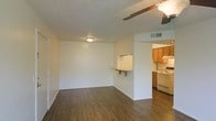 This single-floor space offers a practical and functional layout designed for comfortable everyday living. The main living area is open and airy, featuring dark hardwood-style flooring that extends throughout, creating a cohesive and warm aesthetic. A ceiling fan overhead provides both lighting and ventilation, while a flush-mount light fixture illuminates the room evenly. The textured ceiling adds subtle visual interest to the space.

A pass-through breakfast bar connects the living area to the kitchen, offering a convenient spot for casual dining or additional counter space. The kitchen is well-equipped with warm honey-toned wooden cabinetry, white appliances including a range and refrigerator, and an under-cabinet range hood. The cabinetry provides ample storage, and the layout ensures efficient use of the available space.

The open connection between the living area and kitchen creates an inviting flow, making the space feel larger than it might otherwise appear. Neutral white walls throughout offer a clean, versatile backdrop suitable for a variety of décor styles. A visible HVAC vent in the ceiling suggests central climate control, adding to the overall comfort and functionality of the space.
