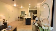 This single-floor residence showcases an open-concept design that fluidly connects the living, dining, and kitchen areas into one cohesive, functional space. The layout maximizes usability while maintaining an airy, welcoming atmosphere throughout.

The kitchen is outfitted with dark espresso cabinetry that provides a striking contrast against the light surroundings. Stainless steel appliances, including a full-size refrigerator, contribute a modern, polished feel. A central island with a built-in sink and bar seating serves as both a practical workspace and a social hub, ideal for casual dining or entertaining.

Adjacent to the kitchen, the dining area comfortably accommodates a mid-century modern table and chairs, enhanced by decorative wall art that adds a touch of personality. Recessed lighting throughout the space ensures consistent, warm illumination, complementing the natural tones of the light hardwood-style flooring that runs seamlessly across the entire unit.

The living area flows naturally from the dining space, grounded by a textured area rug and thoughtfully placed décor. Overall, the space balances contemporary style with everyday practicality.
