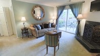 This single-floor residence centers around a thoughtfully designed living room that combines comfort and style. The space is finished with soft, neutral carpeting that adds warmth underfoot, while the walls are painted in a calming sage green hue that creates a tranquil atmosphere. Crown molding details along the ceiling provide an elegant architectural touch.

Natural light is a standout feature, with a full-width sliding glass door that opens directly onto an outdoor patio, where a bistro table and chairs are visible among lush greenery and flowering plants. Sheer and draped blue-grey curtains frame the door, offering both privacy and a sophisticated aesthetic that ties the room's color palette together.

The living room is furnished with a generous taupe sofa accented by decorative pillows in blue and brown tones, accompanied by a striped accent chair and coordinating side tables with warm-toned lamps. A striking ornate gold-framed circular mirror serves as a focal wall piece, enhancing the room's classic, refined character. The overall ambiance is welcoming, balanced, and bathed in natural light.