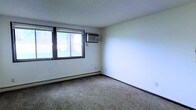 This single-floor space presents a straightforward and functional interior. The room visible in the image features wall-to-wall carpeting in a neutral beige tone, contributing to a quiet and understated atmosphere. White walls keep the space feeling open and bright. A horizontal sliding window along one wall allows natural light to filter in, offering views of surrounding greenery outdoors. A window-mounted air conditioning unit is installed directly beside the window, providing in-unit climate control during warmer months. Along the base of the wall, a baseboard heating system ensures warmth during cooler seasons, indicating year-round comfort. The ceiling has a subtle textured finish, consistent with practical residential construction. The room's simple layout provides a blank canvas adaptable to a variety of uses. Overall, the space reflects a no-frills, functional design with essential amenities in place, making it a practical and comfortable environment.