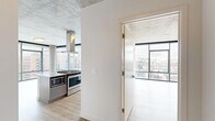 This contemporary urban residence offers a thoughtfully designed single-floor layout with two bedrooms and three bathrooms. The architectural design is defined by exposed concrete ceilings and structural columns, which lend an industrial character balanced by clean white walls and light hardwood flooring throughout.

Floor-to-ceiling windows with sleek black frames are a standout feature, flooding the living spaces with natural light and framing expansive city skyline views. The open living area benefits directly from this wall of glass, creating a bright and airy atmosphere. A prominent concrete column anchors the living space, reinforcing the building's industrial-modern aesthetic.

The kitchen is well-equipped with gray shaker-style cabinetry, white quartz countertops, a hexagonal tile backsplash, and a full suite of stainless steel appliances including a gas range, built-in microwave, dishwasher, and refrigerator. An island provides additional workspace and storage.

The primary bedroom features its own floor-to-ceiling window wall with city views. The bathrooms are finished with gray vanities, white countertops, and tile flooring, maintaining a cohesive, modern palette throughout the entire residence.