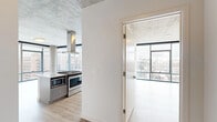 This contemporary urban residence offers a thoughtfully designed single-floor layout with two bedrooms and three bathrooms. The architectural design is defined by exposed concrete ceilings and structural columns, which lend an industrial character balanced by clean white walls and light hardwood flooring throughout.

Floor-to-ceiling windows with sleek black frames are a standout feature, flooding the living spaces with natural light and framing expansive city skyline views. The open living area benefits directly from this wall of glass, creating a bright and airy atmosphere. A prominent concrete column anchors the living space, reinforcing the building's industrial-modern aesthetic.

The kitchen is well-equipped with gray shaker-style cabinetry, white quartz countertops, a hexagonal tile backsplash, and a full suite of stainless steel appliances including a gas range, built-in microwave, dishwasher, and refrigerator. An island provides additional workspace and storage.

The primary bedroom features its own floor-to-ceiling window wall with city views. The bathrooms are finished with gray vanities, white countertops, and tile flooring, maintaining a cohesive, modern palette throughout the entire residence.