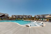 Located at 100 Edison Ave NW in Huntsville, Alabama, this outdoor amenity area showcases a generously sized, freeform resort-style swimming pool as its centerpiece. The pool features a distinctive curved design with built-in in-water lounge chairs that allow residents to relax partially submerged, offering a true resort experience. The surrounding deck is finished in a smooth, light-colored concrete that reflects the abundant Alabama sunshine, and is lined with a large selection of chaise lounges and market umbrellas, providing ample space for sunbathing and leisure.

Adjacent to the pool, a covered cabana with ceiling fans and stone pillar accents offers a shaded retreat, ideal for outdoor gatherings. A separate pergola-style wooden structure provides an additional shaded seating area, adding architectural variety to the space. The pool area is enclosed by a black iron fence, offering both security and open sightlines to the surrounding multi-story residential buildings and lush green lawns beyond. The overall ambiance is bright, open, and resort-inspired, designed to encourage outdoor living under the expansive Alabama sky.