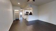 This single-floor space showcases an open-concept design that seamlessly connects the living area to the kitchen. Dark hardwood-style flooring runs throughout, providing a rich contrast against the crisp white walls and cabinetry. The living area is generously proportioned, outfitted with a ceiling fan and recessed lighting that together ensure comfortable illumination at any time of day.

The kitchen features white cabinetry, dark granite-style countertops, and black appliances, including a refrigerator and microwave. A peninsula-style breakfast bar extends into the living space, offering additional seating and defining the boundary between the two areas without sacrificing the open feel.

Adjacent to the main living space, a built-in shelving unit provides practical storage and display options. A doorway leads to a secondary room, which benefits from natural light through a window fitted with vertical blinds. HVAC vents are positioned throughout the ceiling, ensuring consistent climate control. The overall ambiance is clean, modern, and move-in ready, with a neutral palette that adapts easily to a variety of furnishing styles.