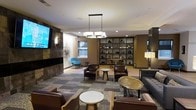 This single-floor lounge space is thoughtfully designed to offer a sophisticated yet comfortable environment. The room is anchored by a large flat-screen television mounted on a sleek, dark stone-clad accent wall, complemented by an eye-catching starburst wall sculpture that adds a mid-century modern touch. A statement pendant chandelier with a rectangular frame hangs centrally, casting warm light across the space and enhancing the inviting atmosphere.

Seating is arranged in multiple conversational groupings, featuring rich brown leather armchairs and sofas paired with marble-top and metallic accent tables. A standout blue accent chair provides a pop of color against the otherwise earthy, neutral palette. The flooring transitions between patterned area rugs and hard surface flooring, helping to define distinct zones within the open layout.

A built-in shelving unit spanning one wall displays a curated collection of decorative objects and books, adding depth and visual interest. Recessed lighting throughout supplements the pendant and floor lamps, ensuring well-distributed illumination. A small dining or café-style seating area is visible in the background, extending the space's functionality. Natural light filters in through windows, contributing to the overall warm and welcoming ambiance.