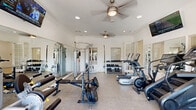 Located at 1101 Longhorn Rd in Fort Worth, TX, this fitness center offers a well-rounded wellness facility spread across a single floor. The space is thoughtfully organized into distinct zones, each serving a specific purpose in the health and fitness journey.

The main exercise room is the heart of the facility, featuring durable speckled flooring, crown molding, and ceiling fans that keep the space comfortable during workouts. Large mirrors line the walls, enhancing the sense of openness. The room is equipped with a comprehensive selection of machines and free weights, including treadmills, an elliptical, a stair climber, a cable machine with TRX straps, a weight bench with plates, and a full dumbbell rack. Wall-mounted TVs provide entertainment, while ample natural light streams in through multiple windows.

A separate room serves as a tanning and relaxation space, featuring a tanning bed bathed in ambient blue lighting, wood-look flooring, a leather daybed, a cowhide area rug, and darkening blinds for privacy. Its bold navy accent wall and equestrian artwork give it a distinctive, lounge-like atmosphere.

The hallway connects the spaces with a warm, welcoming touch — highlighted by light blue walls adorned with decorative air plant arrangements and a console desk seating area, offering a pleasant transition between the active and restorative areas of the facility.