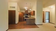 This single-floor space showcases an open-concept layout that connects the kitchen and living areas in a functional and visually cohesive design. The kitchen is a standout feature, highlighted by soaring vaulted ceilings that amplify the sense of space and airiness throughout. Medium-toned wood cabinetry provides ample storage, complemented by a built-in microwave and black appliances, including a gas range. A peninsula breakfast bar extends from the cabinetry, offering additional workspace and a casual dining option. Three pendant lights hang above the kitchen area, providing focused task lighting that enhances the warm ambiance. The flooring transitions from hardwood-style planks in the kitchen to plush carpeting in the adjoining living area, clearly defining each zone while maintaining an open feel. A hallway visible to the left leads to additional rooms, suggesting a practical and well-organized floor plan. The neutral wall tones and warm wood accents throughout contribute to an inviting atmosphere, making this space both comfortable and versatile for everyday living.