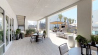 The pool amenity area at 1660 Metro in Chula Vista is a thoughtfully designed outdoor retreat that seamlessly blends covered and open-air spaces. A generously sized covered terrace provides shade for multiple seating arrangements, including casual dining tables with chairs and a dedicated lounge area furnished with contemporary upholstered sofas accented with bold navy and chevron throw pillows. Large planters filled with lush succulents, tropical greenery, and manicured shrubs are positioned throughout, adding vibrant natural texture to the clean, modern aesthetic. Light-toned plank decking runs underfoot, unifying the covered and open sections of the space. Beyond the covered terrace, the open courtyard features a pergola with a warm-toned canopy, a built-in fireplace surround, and additional seating areas complete with market umbrellas for sun protection. Tall, slender palm trees frame the background, reinforcing the Southern California ambiance. A wall-mounted flat-screen television is integrated into the covered lounge area, enhancing the space's functionality for outdoor entertainment. Glass entry doors connect the covered terrace to the surrounding grounds, flooding the interior-facing areas with abundant natural light. The overall design is cohesive and resort-inspired, offering residents a polished and comfortable outdoor living experience.