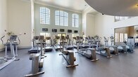 Located at Park Central Drive in Orlando, Florida, this spacious fitness center offers an impressive and well-appointed workout environment spread across a generously sized single-floor layout. The facility is architecturally distinguished by soaring double-height ceilings in the main cardio area, framed by rows of large, arched multi-pane windows that flood the space with natural light and offer views of the surrounding greenery. Crown molding and clean white walls add a polished, upscale aesthetic throughout. The main cardio zone is extensively equipped with treadmills, elliptical trainers, and step machines, all arranged to take full advantage of the bright, airy atmosphere. Multiple wall-mounted televisions are positioned for convenient viewing during workouts. A staircase with decorative metal railings leads to an upper-level mezzanine balcony that overlooks the main floor, adding a dynamic architectural dimension to the space. A dedicated strength training area features a wide selection of selectorized weight machines, free-weight stations, and a dumbbell rack lined against mirrored walls, enhancing the sense of space. Additional windows in this zone provide natural light and outdoor views. The durable rubber flooring throughout ensures both safety and functionality, making this fitness center a comprehensive and inviting facility for all types of training.