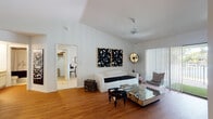 Located at 3101 N 40th Ave in Hollywood, Florida, this single-floor home offers a thoughtfully designed layout with a cohesive modern aesthetic throughout. The living area immediately impresses with its vaulted ceilings and warm hardwood flooring, creating an open and airy atmosphere. A striking pair of black-and-white abstract artworks anchors the main wall, complemented by a white sofa, a mirrored glass coffee table, and eclectic accent pieces. A ceiling fan keeps the space comfortable, while a sliding glass door with vertical blinds opens to an outdoor patio area, allowing natural light to flow freely into the room. A wall-mounted television and additional seating complete the living space. The galley-style kitchen features light wood cabinetry lining both sides, a double-basin stainless steel sink, and full appliances including a refrigerator and range, with a pass-through hallway connecting to the rest of the home. The bedroom is boldly styled with a dark charcoal accent wall serving as a dramatic backdrop, paired with a desk area and warm wood-tone flooring. The bathroom offers a wide vanity with an oversized mirror, extended countertop space, and a shower-tub combination, all finished in a clean, bright palette.