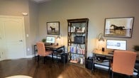 Located in Olde Towne, this single-floor space offers a thoughtfully designed interior with a distinctive blend of industrial and rustic aesthetics. The office area is a standout feature, showcasing rich, dark hardwood flooring that anchors the room with warmth and character. Two industrial-style pipe-frame desks provide ample workspace, each accompanied by task lighting from gooseneck-style desk lamps that cast a warm, focused glow throughout the room. A tall, rustic wooden bookshelf with cross-brace detailing serves as a central organizing element, filled with an extensive collection of books and topped with a printer, blending practicality with visual appeal. Framed artwork, including a classic equestrian portrait, adorns the soft, neutral-toned walls, adding a refined touch to the overall atmosphere. The space is designed to accommodate multiple users comfortably, making it well-suited for collaborative or individual productivity. The cohesive use of dark wood tones, metal accents, and warm lighting creates an inviting and professional ambiance that is both functional and aesthetically engaging.