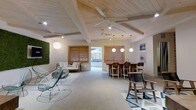 This single-floor space makes a strong architectural impression with its vaulted ceiling clad in light pine wood and accented by exposed beams, creating an airy, resort-like atmosphere. Ceiling fans and recessed lighting work alongside globe pendant lights to illuminate the open layout effectively. The space is divided into distinct functional zones while maintaining an open, connected feel. A lounge area toward one end features contemporary seating, including a wire-frame accent chair and a low-profile sofa, complemented by a living moss wall installation that adds a natural, organic element to the interior. A dining or bar area anchors the central portion of the space, featuring a vibrant countertop surface surrounded by tall wooden stools and pendant lighting overhead. Louvered wooden windows frame views of an outdoor area, allowing natural light and ventilation to filter through while maintaining a sense of privacy. The flooring transitions from polished concrete to a geometric-patterned area rug, adding visual interest underfoot. The overall ambiance is clean, modern, and relaxed, with a coastal-contemporary aesthetic defined by natural materials, neutral tones, and carefully curated furnishings.
