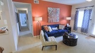This single-floor residence makes a striking first impression with its thoughtfully curated interior design. The living room serves as the centerpiece of the home, showcasing a bold terracotta-orange accent wall that anchors the space with warmth and energy. A large piece of abstract wall art complements the color palette, adding an organic, nature-inspired focal point above the seating area. The living area is furnished with a rich navy blue sofa adorned with layered throw pillows in coordinating earth tones, paired with a tufted accent chair featuring a botanical print. Two sleek floor lamps with white shades provide warm ambient lighting, while a dark round coffee table grounds the seating arrangement. The neutral wall-to-wall carpeting offers a soft contrast to the vibrant accent wall, contributing to a balanced and cohesive aesthetic. A hallway visible from the living area leads deeper into the home, hinting at additional rooms and functional spaces beyond. The open sightlines and well-proportioned layout lend the interior a spacious, airy quality. Overall, the space blends bold color with comfortable furnishings to create a home that feels both stylish and livable.