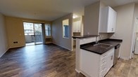 This single-floor residence offers a thoughtfully designed open-concept layout that seamlessly connects the kitchen and living area. Rich, dark hardwood-style flooring runs throughout, adding warmth and a sense of continuity across the space. The kitchen is outfitted with crisp white cabinetry, dark countertops, and a double-basin sink, creating a clean, modern aesthetic. A pass-through window connects the kitchen to an adjacent area, enhancing the open feel and allowing natural light to flow freely. The living area is spacious and inviting, anchored by a large sliding glass door that opens onto a private balcony, bringing in abundant natural light and views of the surrounding exterior. The balcony is enclosed with a slatted privacy screen, offering a comfortable outdoor retreat. Neutral gray walls provide a versatile backdrop throughout the interior, complementing both the light cabinetry and the deeper tones of the flooring. The overall ambiance is clean, bright, and functional, making excellent use of the available space while maintaining a welcoming and livable atmosphere suited for everyday comfort.