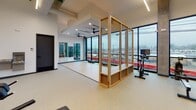 Located on W Corporation Street in the heart of Durham, NC, this contemporary fitness center spans two floors and is thoughtfully designed to support a wide range of workout styles and routines. The facility features a modern industrial aesthetic, with exposed concrete columns, high ceilings, and sleek linear lighting that runs throughout the space, creating a clean and energizing atmosphere.

The main workout floor is generously proportioned and equipped with a full array of strength and cardio machines, including treadmills, stair climbers, ellipticals, cable machines, and free weight benches with dumbbell racks. Floor-to-ceiling curtain wall windows line the perimeter, flooding the space with natural light and offering open views of the surrounding Durham cityscape — an especially motivating backdrop during workouts.

A dedicated studio or flex area is defined by a light wood-framed glass partition, featuring hardwood-style flooring, wall-to-wall mirrors, and built-in shelving — ideal for stretching, yoga, or functional training. Adjacent to the main floor is a well-appointed amenity station with custom wood cabinetry, open shelving, and a counter workspace.

Ceiling fans are installed throughout to enhance airflow and comfort. The overall design balances industrial character with warm wood accents, creating a welcoming and fully functional fitness environment suited for daily use.
