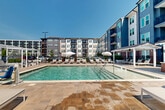 Located at 235 Verbena St in Charlotte, NC, this outdoor amenity space delivers a resort-style experience centered around two generously sized swimming pools. The primary pool features sparkling blue-green water, water jet features along the entry steps, and is framed by a broad stone-tiled sun deck lined with rows of chaise lounges and market umbrellas — ideal for relaxing under Charlotte's sunny skies. A secondary pool area is anchored by a brick-accented clubhouse pavilion with a pitched roof, providing a shaded retreat alongside additional lounge seating and umbrella stations.

A standout feature of the space is the elevated pergola lounge area, outfitted with slatted white-framed shade structures, built-in planters, and cushioned chaise lounges — creating a sophisticated sunbathing and socializing zone with open views of the surrounding neighborhood. The overall design blends contemporary and traditional architectural elements, with the multi-story residential buildings — finished in contrasting blue, gray, and brick tones — forming an impressive backdrop that enhances the sense of community. The stone-paved decking throughout ties the areas together cohesively, while thoughtful landscaping adds greenery and texture to the open-air environment.