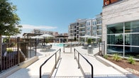 Located at 2005 W Campbell Rd in Garland, Texas, this single-story amenity space is part of a contemporary multi-story residential community defined by clean architectural lines, neutral stucco and stone exteriors, and expansive balconies throughout the surrounding building facade.

The centerpiece of the outdoor area is a large, resort-style swimming pool with crystal-clear blue water. The pool deck is generously proportioned, featuring multiple tanning ledges with built-in seating elements, lounge chairs, and umbrella-shaded dining tables arranged across the open hardscape. A sleek pergola structure anchors one end of the courtyard, providing a shaded gathering zone.

Adjacent to the pool area, a fully equipped outdoor kitchen sits beneath a modern louvered pergola roof with exposed steel framing. The cooking station includes multiple stainless steel grills set into stone-clad counters, offering a functional and stylish space for outdoor entertaining.

A covered patio lounge connects directly to the pool courtyard through large glass doors and floor-to-ceiling windows. This sheltered space is finished with light tile flooring and furnished with wicker seating in teal and neutral tones, creating a comfortable transitional area between indoors and the pool environment. Iron fencing and manicured landscaping complete the well-appointed outdoor setting.