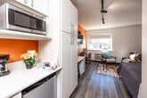 Located at 4117 University Ave in Cedar Falls, Iowa, this efficiently designed studio unit makes smart use of its compact footprint across a single floor. The open-concept layout seamlessly integrates the kitchen, living, and sleeping areas into one cohesive space. A bold orange accent wall serves as a striking focal point, adding warmth and energy to the interior, while light wood-style flooring and white walls keep the overall aesthetic bright and clean.

The kitchenette is thoughtfully arranged with white cabinetry extending to the ceiling for ample storage, a built-in microwave, a compact refrigerator, a sink, and counter space — all the essentials for everyday meal preparation. Track lighting runs along the textured ceiling, providing flexible illumination throughout the unit.

The main living area features a wall-mounted flat-screen TV, a dedicated study desk with seating, and a comfortable lounge chair and sofa — ideal for both relaxation and productivity. A space-saving Murphy bed folds down to reveal a full-size sleeping surface, complemented by colorful abstract wall art above. A vibrant multicolored area rug anchors the room and adds a playful design touch. A window allows natural light to filter in, offering views of the surrounding area.

The private bathroom includes a walk-in shower with a curtain enclosure, a toilet, and consistent orange accent detailing that ties the space together.