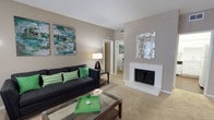 This single-story space centers around a well-appointed living room that combines comfort with contemporary style. The living area is finished with wall-to-wall carpeting in a warm beige tone, contributing to an inviting and cozy atmosphere throughout. Neutral taupe walls provide a versatile backdrop that complements both the furnishings and decorative elements.

A standout feature of the living room is the white-painted brick fireplace, which serves as a natural focal point along one wall. Above it, a geometric mirrored wall accent adds a modern, reflective touch that enhances the sense of light in the room. On the opposite wall, bold abstract artwork in shades of green and teal introduces a vibrant pop of color that ties together the room's décor scheme.

The space is furnished with a classic dark sofa accented with green chevron and solid throw pillows, paired with a glass-topped coffee table that keeps the area feeling open and uncluttered. A floor lamp provides warm ambient lighting. A hallway visible from the living area leads to additional rooms, suggesting a practical and connected layout. Overall, the space presents a clean, modern aesthetic with welcoming, residential warmth.