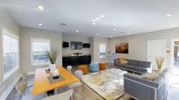 This single-floor community space is designed with an open-concept layout that seamlessly blends multiple functional zones into one cohesive, welcoming environment. The dining area features a warm orange wooden table paired with contemporary white chairs, ideal for casual meals or group gatherings. Adjacent to it, a kitchenette with dark cabinetry and granite-style countertops provides a convenient refreshment station, complemented by a wall-mounted flat-screen television above.

The lounge area is anchored by a plush gray sectional sofa adorned with gold accent pillows, centered around a sleek glass coffee table resting atop a decorative area rug. Colorful ottomans in blue and orange add a playful, modern touch throughout the space, encouraging flexible seating arrangements. A high-top bar table with stools near the window offers an additional spot for conversation or casual work.

Natural light filters in through multiple windows positioned along the walls, brightening the light gray painted interior and wood-look flooring that runs throughout. Recessed ceiling lighting further enhances the clean, airy feel of the space. A framed cityscape artwork adds a decorative focal point to the lounge section. Overall, the space exudes a contemporary, energetic ambiance suited for socializing and community interaction.