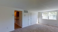 Located at 6537 D Falkirk Rd in Baltimore, MD 21239, this single-floor residence offers a straightforward and functional layout designed for comfortable everyday living. The main living area features wall-to-wall carpeting, creating a warm and inviting atmosphere, while a large window allows natural light to fill the space and provide views of the surrounding exterior. Clean white walls and neutral tones throughout contribute to a bright and open feel. A centrally positioned HVAC vent ensures climate control across the living space. Adjacent to the main living area, the kitchen is accessible through an open doorway and is outfitted with wooden cabinetry, a dark countertop, and tile flooring, offering a functional cooking and preparation space. Louvered closet doors provide convenient storage solutions within the living area, adding to the unit's practicality. The overall design is clean and utilitarian, making it well-suited for those seeking a low-maintenance, move-in-ready space. The single-floor configuration ensures easy accessibility throughout, while the combination of carpeted living spaces and tiled kitchen areas provides a clear and logical separation of functional zones within the home.