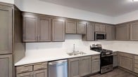 This single-story space showcases a well-appointed kitchen that serves as a functional centerpiece. The kitchen features full-height upper and lower shaker-style cabinetry finished in a warm taupe-gray tone, providing generous storage throughout. Sleek brushed nickel hardware accents each cabinet door and drawer, contributing to a cohesive, contemporary aesthetic. The crisp white countertops offer ample workspace and contrast neatly against the darker cabinetry. Stainless steel appliances include a gas range with multiple burners, an over-the-range microwave, and a dishwasher, equipping the kitchen for practical everyday use. A stainless steel double-basin sink is centrally positioned along the counter with a simple modern faucet. The white walls and neutral color palette throughout create a clean, bright atmosphere. The cabinetry extends along the full length of the wall, maximizing storage capacity, while the overall layout supports efficient workflow. The space maintains a straightforward, modern design sensibility with durable, low-maintenance finishes suited for everyday living.