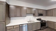 This single-story space showcases a well-appointed kitchen that serves as a functional centerpiece. The kitchen features full-height upper and lower shaker-style cabinetry finished in a warm taupe-gray tone, providing generous storage throughout. Sleek brushed nickel hardware accents each cabinet door and drawer, contributing to a cohesive, contemporary aesthetic. The crisp white countertops offer ample workspace and contrast neatly against the darker cabinetry. Stainless steel appliances include a gas range with multiple burners, an over-the-range microwave, and a dishwasher, equipping the kitchen for practical everyday use. A stainless steel double-basin sink is centrally positioned along the counter with a simple modern faucet. The white walls and neutral color palette throughout create a clean, bright atmosphere. The cabinetry extends along the full length of the wall, maximizing storage capacity, while the overall layout supports efficient workflow. The space maintains a straightforward, modern design sensibility with durable, low-maintenance finishes suited for everyday living.