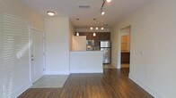 This single-floor unit showcases an open-concept design that seamlessly connects the living area to the kitchen. The flooring throughout the main living space consists of rich, dark hardwood-style planks that add warmth and visual depth to the interior.

The kitchen is positioned at the heart of the unit, featuring a raised breakfast bar that doubles as a partition between the living and cooking areas. Dark espresso-toned cabinetry provides ample storage and contrasts cleanly against the light countertops and white walls. Stainless steel appliances, including a refrigerator, microwave, and range, contribute a modern, polished aesthetic. Pendant lighting hangs above the kitchen area, complementing the track lighting installed on the ceiling of the main living space.

A hallway visible from the main living area leads to additional rooms, with a glimpse of a secondary space that appears to include a sink and cabinetry, suggesting a bathroom or utility area. The overall layout is practical and efficient, with a neutral color palette of white walls and dark accents creating a clean, contemporary ambiance throughout the space.