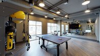 This single-floor fitness and recreation facility offers a well-rounded environment designed to support a variety of physical activities and leisure. The space features an open, industrial-style aesthetic, with exposed ductwork and conduit along the ceiling, complemented by warm pendant lighting that illuminates the hardwood-style flooring below.

At the center of the space sits a full-size ping-pong table, making it a natural social hub for recreational play. Along one wall, TRX suspension training straps and a stability ball are mounted and ready for use, providing a dedicated zone for functional fitness training. A row of stationary cycling bikes lines the perimeter, ideal for group or individual cardio sessions.

A large wall-mounted flat-screen display adds a multimedia element to the space, suitable for streaming workout content or entertainment. Full-length mirrors on one wall enhance the sense of openness and are practical for form-checking during exercise. Large windows along the exterior wall flood the interior with natural light, creating an energizing atmosphere throughout.

The overall ambiance is modern and purposeful, balancing the raw character of its industrial design with well-organized fitness amenities, making it a versatile space suited for both active workouts and casual recreation.