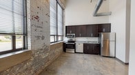 This 1-bedroom, 1-bathroom flat delivers a compelling blend of industrial character and modern finishes across a single open floor. The living and kitchen area immediately impresses with soaring ceilings that amplify the sense of space, exposed ductwork overhead, and a striking wall of original exposed brick that adds raw, historic texture. Large windows with horizontal blinds flood the open-plan area with natural light while offering street-level views. Polished concrete floors run throughout, tying the industrial aesthetic together cohesively.

The kitchen is well-appointed with dark espresso cabinetry extending to the upper reaches of the tall walls, granite countertops, and a full suite of stainless steel appliances including a refrigerator, electric range, microwave, and dishwasher. A single-basin undermount sink completes the functional layout.

The bedroom is a calm, neutral space with clean white walls, concrete flooring, and ample room for furnishings. The bathroom features a matching dark vanity with granite countertop and an undermount sink, a large mirror, and a full tub-shower combination, maintaining the consistent design language found throughout the flat. This space is ideal for those who appreciate urban industrial style with modern conveniences.