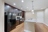 This single-floor residence offers a thoughtfully designed layout that balances modern finishes with comfortable living. The kitchen serves as a central highlight, showcasing rich dark espresso cabinetry that extends to the ceiling for ample storage, granite countertops, a tile backsplash, and a large center island with a built-in sink. Pendant lighting illuminates the island, while recessed ceiling lights brighten the full kitchen space. Black appliances, including a refrigerator with an ice and water dispenser, electric range, and microwave, complete the culinary setup. Warm hardwood flooring flows throughout the main living and common areas, lending a cohesive, inviting feel. Living spaces are generously proportioned, with neutral warm-toned walls and ceiling fans that enhance comfort. Multiple windows dressed with horizontal blinds allow natural light to filter in while offering views of surrounding greenery. One room opens directly onto a private balcony, further connecting the interior to the outdoors. The bathroom features large-format tile flooring, a vanity with granite countertop and dark cabinetry, a glass-enclosed shower with tile surround, and a window for natural light. A spacious walk-in closet with wire shelving and carpet flooring provides excellent storage capacity, rounding out this well-equipped residence.