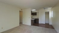 This single-floor residential unit offers a practical and comfortable layout designed for everyday living. The main living area is spacious and carpeted, providing a warm and inviting atmosphere. A ceiling fan overhead ensures year-round comfort, while neutral-toned walls create a clean, versatile backdrop throughout the space.

The living area opens directly into the kitchen, which features dark wood cabinetry, a smooth countertop surface, and a peninsula-style breakfast bar that doubles as a casual dining or workspace. The kitchen is finished with hard flooring, contrasting with the soft carpet of the living area. A skylight above the kitchen brings in natural light, brightening the cooking space. The kitchen also includes a full-size refrigerator and ample under-counter storage.

A hallway visible from the living area leads to additional rooms, suggesting a well-organized floor plan with clearly separated living and private spaces. The textured ceilings and recessed ventilation throughout reflect a functional, no-frills interior design. Overall, the unit presents a straightforward and efficient living environment suited for those seeking a comfortable, move-in-ready space.
