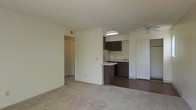 This single-floor residential unit offers a practical and comfortable layout designed for everyday living. The main living area is spacious and carpeted, providing a warm and inviting atmosphere. A ceiling fan overhead ensures year-round comfort, while neutral-toned walls create a clean, versatile backdrop throughout the space.

The living area opens directly into the kitchen, which features dark wood cabinetry, a smooth countertop surface, and a peninsula-style breakfast bar that doubles as a casual dining or workspace. The kitchen is finished with hard flooring, contrasting with the soft carpet of the living area. A skylight above the kitchen brings in natural light, brightening the cooking space. The kitchen also includes a full-size refrigerator and ample under-counter storage.

A hallway visible from the living area leads to additional rooms, suggesting a well-organized floor plan with clearly separated living and private spaces. The textured ceilings and recessed ventilation throughout reflect a functional, no-frills interior design. Overall, the unit presents a straightforward and efficient living environment suited for those seeking a comfortable, move-in-ready space.