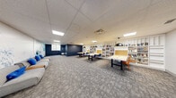 This single-floor collaborative workspace offers a thoughtfully designed environment that blends productivity and comfort. The main area is anchored by several large, light-colored work tables accompanied by blue and orange chairs, providing ample seating for group study or collaborative projects. Striking black dome pendant lights hang above the tables, casting focused illumination across the workspace.

Lining the walls are extensive floor-to-ceiling white built-in bookshelves, densely stocked with a diverse collection of books and reference materials, lending the space an intellectual and resourceful character. Cork board panels integrated into the shelving units offer additional functional space for notes and displays.

The lounge area provides a relaxed retreat, featuring a modern grey sofa with blue accent pillows set against a dramatic navy blue accent wall and a vivid large-scale mountain mural rendered in shades of blue and white, creating a visually dynamic focal point. The same mural extends into the adjoining open area, reinforcing a cohesive aesthetic theme throughout the space.

Underfoot, patterned grey carpet tiles add texture and a professional finish, while a drop ceiling with recessed and flat-panel skylights ensures consistent, even lighting throughout. The overall ambiance is professional yet welcoming, making this space well-suited for learning, collaboration, and creative work.