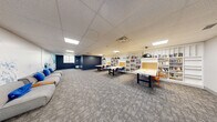 This single-floor collaborative workspace offers a thoughtfully designed environment that blends productivity and comfort. The main area is anchored by several large, light-colored work tables accompanied by blue and orange chairs, providing ample seating for group study or collaborative projects. Striking black dome pendant lights hang above the tables, casting focused illumination across the workspace.

Lining the walls are extensive floor-to-ceiling white built-in bookshelves, densely stocked with a diverse collection of books and reference materials, lending the space an intellectual and resourceful character. Cork board panels integrated into the shelving units offer additional functional space for notes and displays.

The lounge area provides a relaxed retreat, featuring a modern grey sofa with blue accent pillows set against a dramatic navy blue accent wall and a vivid large-scale mountain mural rendered in shades of blue and white, creating a visually dynamic focal point. The same mural extends into the adjoining open area, reinforcing a cohesive aesthetic theme throughout the space.

Underfoot, patterned grey carpet tiles add texture and a professional finish, while a drop ceiling with recessed and flat-panel skylights ensures consistent, even lighting throughout. The overall ambiance is professional yet welcoming, making this space well-suited for learning, collaboration, and creative work.