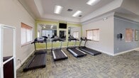 The Weston Place Fitness Center is a thoughtfully designed, single-floor facility offering a comprehensive range of workout amenities. The interior is characterized by a vibrant color scheme of lime green and slate blue accent walls, complemented by neutral flooring with patterned carpet in the exercise areas and smooth tile in transitional spaces. Recessed lighting and flush panel ceiling fixtures ensure the space is well-lit throughout.

The main workout area is generously proportioned and houses an array of cardio equipment, including multiple treadmills by TRUE Fitness, elliptical trainers, and additional cardio machines. A large wall mirror runs along one side of the cardio zone, amplifying the sense of space and allowing exercisers to monitor their form. A wall-mounted television is positioned for convenient viewing during workouts. Multiple windows fitted with wood-slatted blinds allow natural light to filter in.

Adjacent to the main floor, glass-enclosed spaces include a racquetball court with a hardwood floor and standard court markings, as well as a separate multi-purpose room accessible through elegant French doors. This additional room provides flexible space for group activities or alternative training. The facility's open layout, bright finishes, and diverse equipment selection create an energizing and functional environment suited to a wide range of fitness goals.