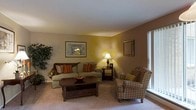 This single-floor space presents a traditionally styled living area designed for comfort and functionality. The living room is anchored by a plush sofa adorned with decorative throw pillows in warm earth tones, complemented by a classic plaid upholstered armchair positioned near a large window that allows natural light to brighten the space. A dark wood coffee table with drawer storage serves as a functional centerpiece, while a matching side table holds a table lamp that casts a warm, ambient glow throughout the room. The walls are painted in a muted warm tone, contributing to the inviting atmosphere. Framed artwork decorates the walls, adding visual interest and a personal touch to the space. Carpeted flooring runs throughout, enhancing the overall sense of warmth and comfort. A lush indoor plant in the corner introduces a natural element to the room. The layout is well-organized, offering a relaxed and functional arrangement that makes efficient use of the available space, suitable for everyday living and casual gatherings.