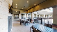 This residential community space is composed of two distinct functional areas designed to serve residents with convenience and comfort. The mail room is a clean, well-lit space finished with light wood-look flooring, neutral walls, and recessed ceiling lights. Rows of individual gray metal mailboxes line multiple walls, providing ample mail and parcel storage for a large number of residents. A central white countertop island offers a practical surface for sorting mail and packages. Security is supported by visible surveillance cameras, and a fire extinguisher station is mounted nearby for safety compliance. A glass-paneled door connects the mail room to a carpeted corridor, offering a glimpse into the broader building interior. The covered outdoor lounge area features a rich wood-paneled ceiling with recessed lighting and a ceiling fan, creating a comfortable shaded retreat. Sturdy square columns anchor the structure, framing open views of the surrounding community through black metal railings. The space is furnished with dark metal dining tables and chairs, ideal for outdoor gatherings, along with a built-in outdoor grill station for resident use. Together, these areas reflect a community-focused design that balances practicality with inviting shared amenities.
