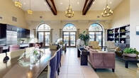 Located at 2615 E Date Palm Street in Ontario, CA, this single-floor space offers an impressive open-plan layout combining a fully equipped kitchen and expansive living area. The space is defined by its dramatic soaring ceilings adorned with rich, dark-stained exposed wood beams, lending a warm yet grand architectural character throughout.

The living area is bright and welcoming, bathed in natural light that streams through a series of large arched windows and French doors framed with elegant drapery. These windows provide views of the surrounding exterior while enhancing the airy, open ambiance. Multiple decorative chandeliers illuminate the space, complementing the warm neutral wall tones and large-format light tile flooring. Built-in dark wood shelving units flank a fireplace, creating an inviting focal point, while lush indoor plants add a natural, refreshing element.

The kitchen is outfitted with rich dark wood cabinetry, gleaming white countertops, and a striking green mosaic tile backsplash. Stainless steel appliances — including a built-in double oven, a large refrigerator, a dishwasher, and a cooktop island — speak to both functionality and refined style. A spacious island with seating provides a casual gathering spot. Together, the kitchen and living areas form a cohesive, elegantly designed space ideal for both everyday use and entertaining.