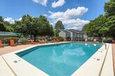 This outdoor pool area in Macon, Georgia, offers a well-appointed recreational space designed for relaxation and leisure. The centerpiece is a generously sized rectangular swimming pool with clear blue water, framed by a broad, paved sundeck that provides ample room for lounging and socializing. The deck is furnished with multiple orange lounge chairs and seating arrangements, allowing guests to comfortably enjoy the sunny Georgia climate.

A single-story poolside building anchors one end of the space, featuring a neutral gray exterior with white-trimmed windows and French doors that open directly onto the pool deck. This structure likely serves as a clubhouse or amenity facility, providing convenient access and a functional backdrop to the outdoor area.

The pool area is enclosed by a black iron fence, offering both security and an open feel, while mature trees and manicured shrubs line the perimeter, providing natural shade and a lush, green backdrop. A shade sail positioned at one end of the deck offers an additional shaded retreat. The combination of open sky, natural greenery, and well-maintained furnishings creates an inviting and comfortable outdoor environment suited to warm-weather enjoyment.