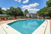 This outdoor pool area in Macon, Georgia, offers a well-appointed recreational space designed for relaxation and leisure. The centerpiece is a generously sized rectangular swimming pool with clear blue water, framed by a broad, paved sundeck that provides ample room for lounging and socializing. The deck is furnished with multiple orange lounge chairs and seating arrangements, allowing guests to comfortably enjoy the sunny Georgia climate.

A single-story poolside building anchors one end of the space, featuring a neutral gray exterior with white-trimmed windows and French doors that open directly onto the pool deck. This structure likely serves as a clubhouse or amenity facility, providing convenient access and a functional backdrop to the outdoor area.

The pool area is enclosed by a black iron fence, offering both security and an open feel, while mature trees and manicured shrubs line the perimeter, providing natural shade and a lush, green backdrop. A shade sail positioned at one end of the deck offers an additional shaded retreat. The combination of open sky, natural greenery, and well-maintained furnishings creates an inviting and comfortable outdoor environment suited to warm-weather enjoyment.