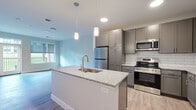 This well-designed one-bedroom, one-bathroom unit in Rochester, MN offers a thoughtful, efficient layout across a single floor. The open-concept living and kitchen area forms the heart of the space, featuring light-toned luxury vinyl plank flooring that extends throughout the main areas. The kitchen is a standout, showcasing gray shaker-style cabinetry with sleek hardware, a classic white subway tile backsplash, granite countertops, and a central island with a built-in sink and pendant lighting overhead. Stainless steel appliances, including a refrigerator, range, and microwave, complete the modern culinary space. The living area benefits from natural light through large windows and a glass door that opens to an outdoor area with views of green surroundings. The bedroom offers plush carpeting, a window with outdoor views, and access to two closets for ample storage. The bathroom is finished with wood-look flooring and chrome fixtures. Additional conveniences include a dedicated laundry area and utility room. Neutral wall tones and clean white trim throughout give the unit a bright, contemporary feel that is both functional and inviting.