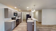 Located at 1100 W 14th St in Webb City, Missouri, this single-floor unit offers a thoughtfully designed, modern living space with a clean, contemporary aesthetic throughout. Light-toned luxury vinyl plank flooring flows seamlessly across the entire unit, creating a cohesive and easy-to-maintain foundation.

The kitchen serves as the centerpiece of the home, featuring gray shaker-style cabinetry, light countertops, and a full suite of black appliances including a range, microwave, and refrigerator. A central island with a built-in sink and pendant lighting overhead provides additional prep space and a natural gathering point. Recessed lighting throughout the kitchen ensures the area is bright and functional.

The open layout connects the kitchen to a generous living area, offering a flexible and airy feel. A wide hallway leads to the bedroom spaces, each featuring ceiling fans, natural light from multiple windows, and direct access to well-designed bathrooms. The primary bathroom is particularly spacious, offering a dual-sink vanity with ample counter space, a tub/shower combination, and direct access to a walk-in closet with built-in shelving. A secondary bathroom mirrors the clean, modern finishes with a single-sink vanity and tub/shower combo. The overall ambiance is fresh, neutral, and move-in ready.